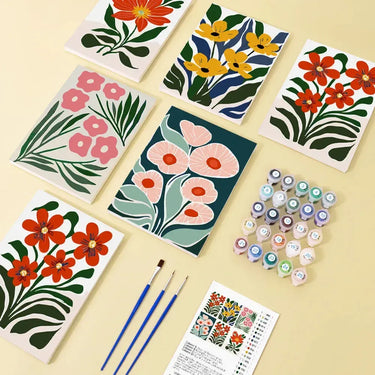 Kukka-aiheinen numeromaalaussetti, jossa kuusi valmista 20x15 cm maalausta, maalipurkit, kolme pensseliä ja ohjeet aseteltuna pöydälle.